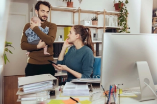 Ein junges Paar mit Kind am Reden, während die Frau im Homeoffice arbeitet.